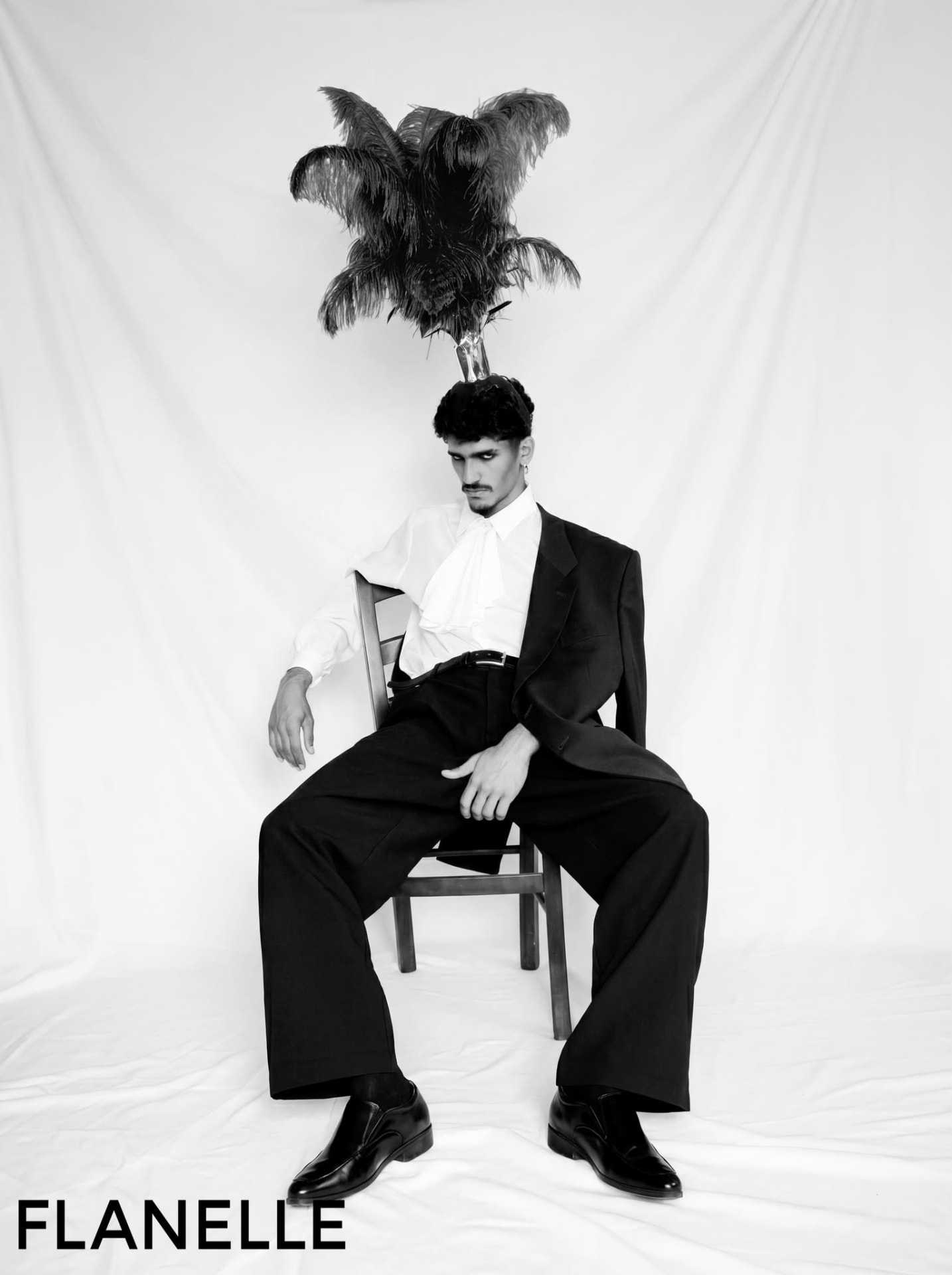 Seated figure in a black suit beneath a black feather headpiece, shadow cast on the backdrop