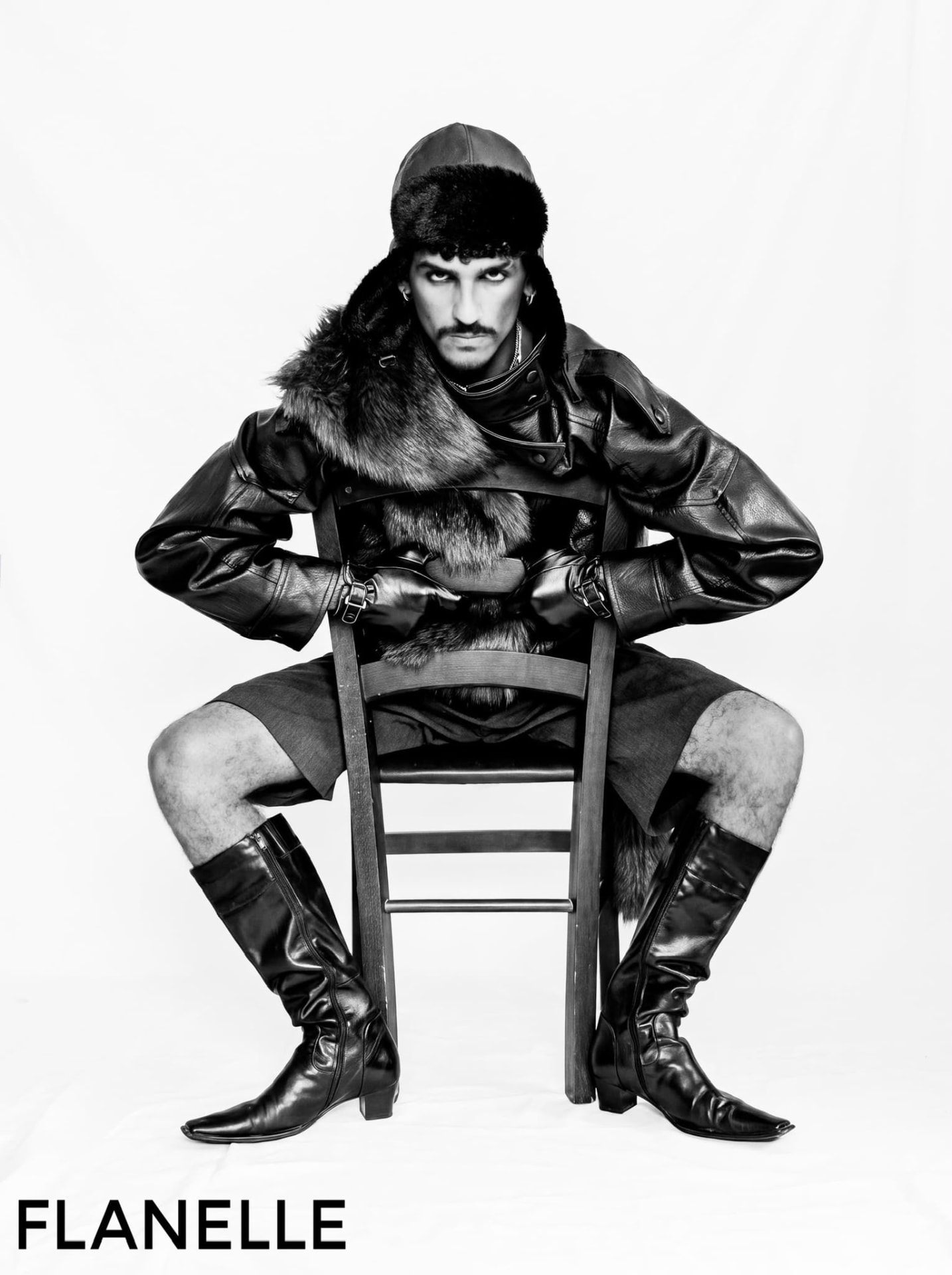 Seated figure in an oversized leather jacket with fur stole, leather cap and knee-high boots